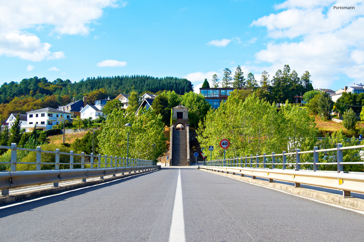 Road leading to a small bridge with railings, a stone monument ahead, and houses on a green hillside.