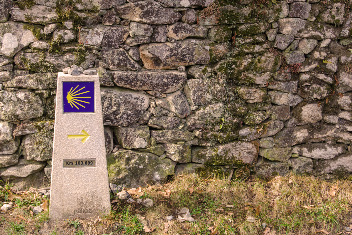 Stone Camino de Santiago marker with yellow shell and arrow beside a rough stone wall.