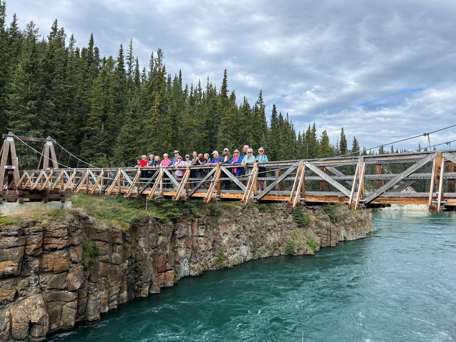 Group of people standing on a wooden bridge over a turquoise river, surrounded by trees and a cloudy sky.
