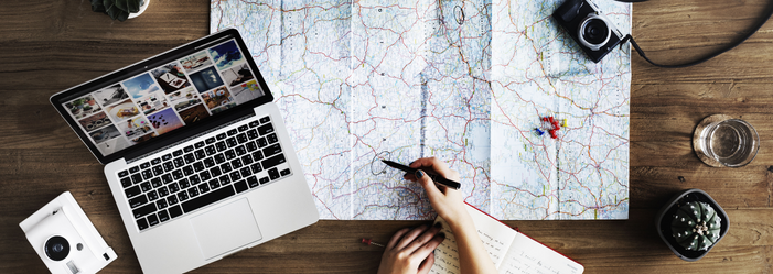 Person planning a trip, using a map and laptop on a wooden desk, with a camera and other travel items.