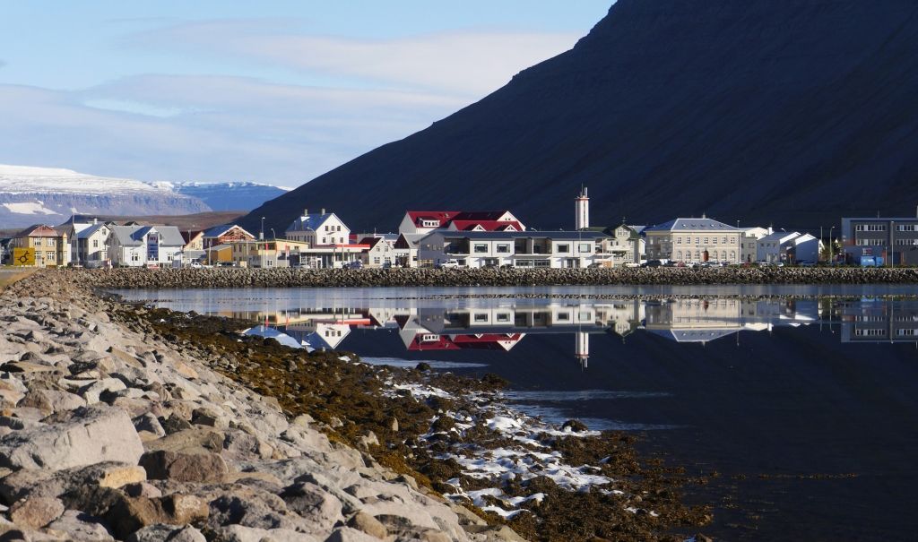 Waterfront town reflects in calm water, framed by dark mountain and rocky shore.