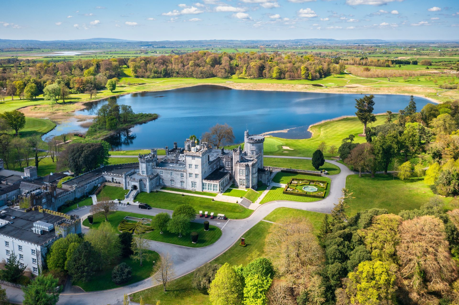 Castle and lake in a green landscape. Gray stone castle with towers, blue lake, and trees under a sunny sky.