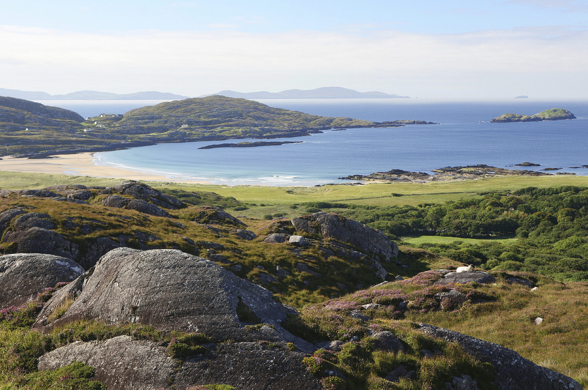 Rocky foreground overlooks a beach, bay, and hills under a blue sky.