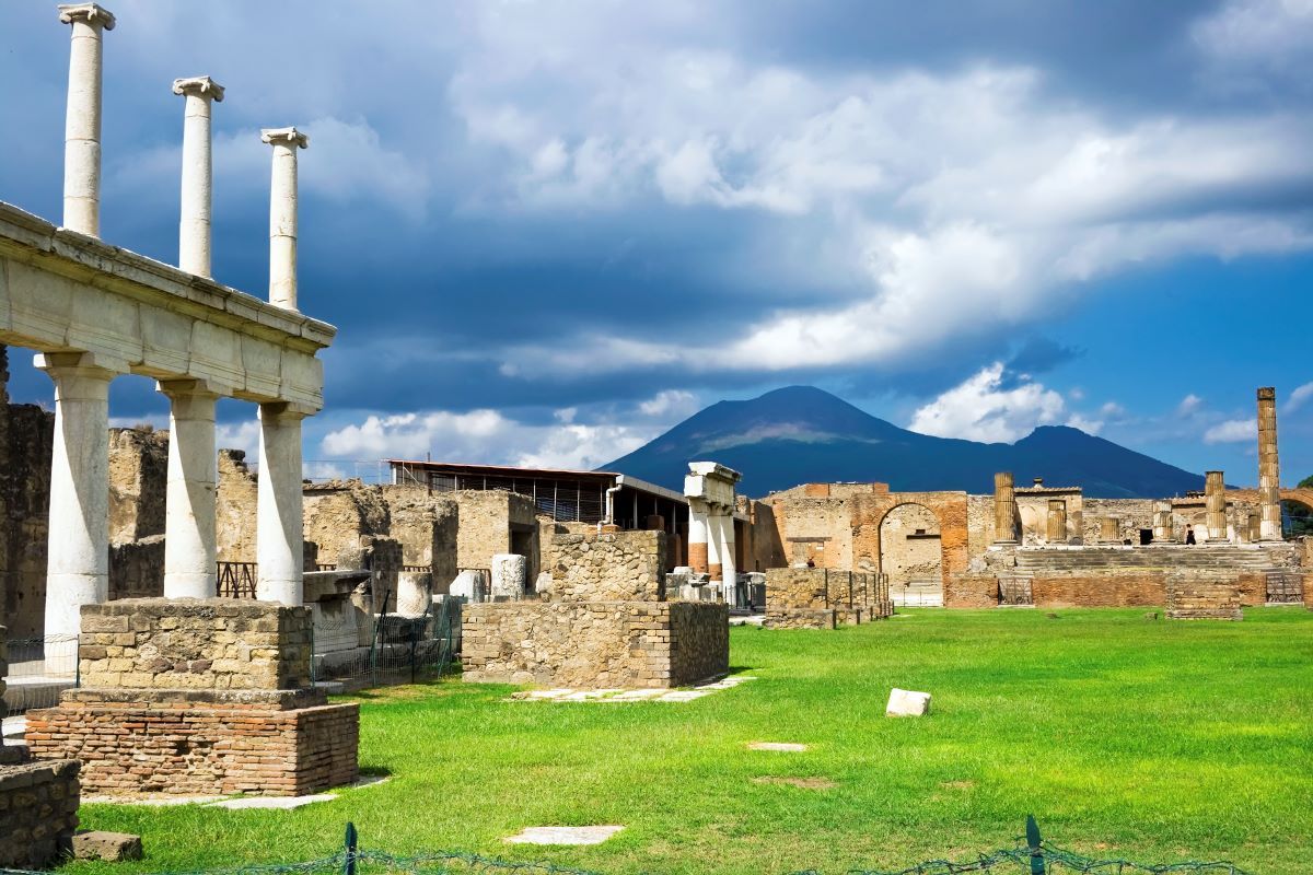Ancient Roman ruins with columns and a grassy foreground, set against mountains and a cloudy sky
