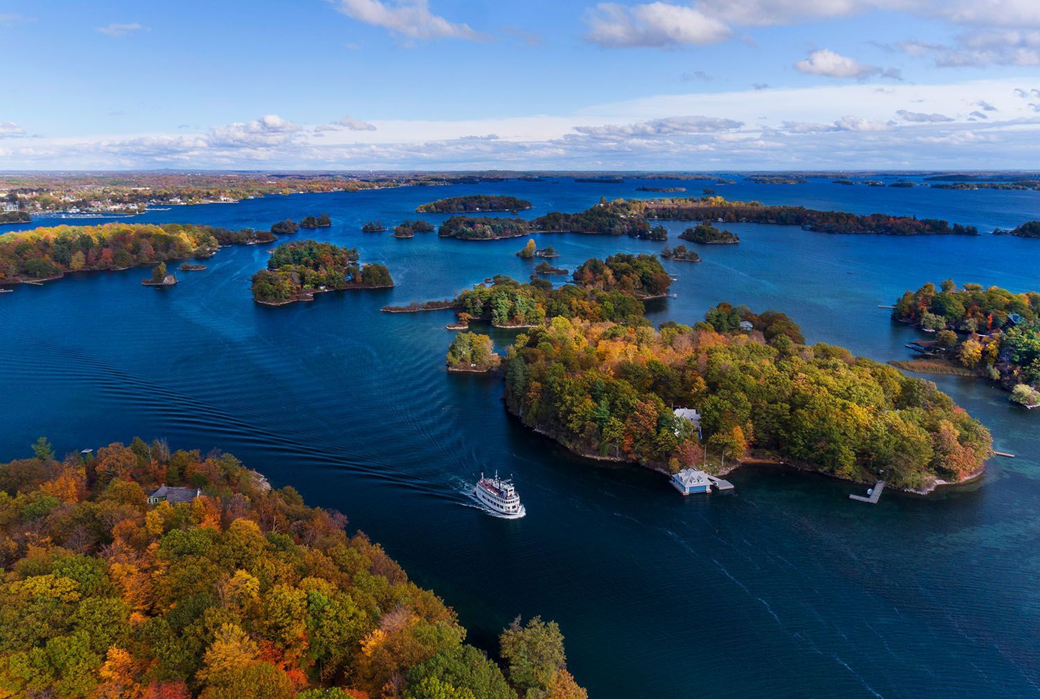 Aerial view of blue water and many small green islands under a bright sky, with a boat cruising through