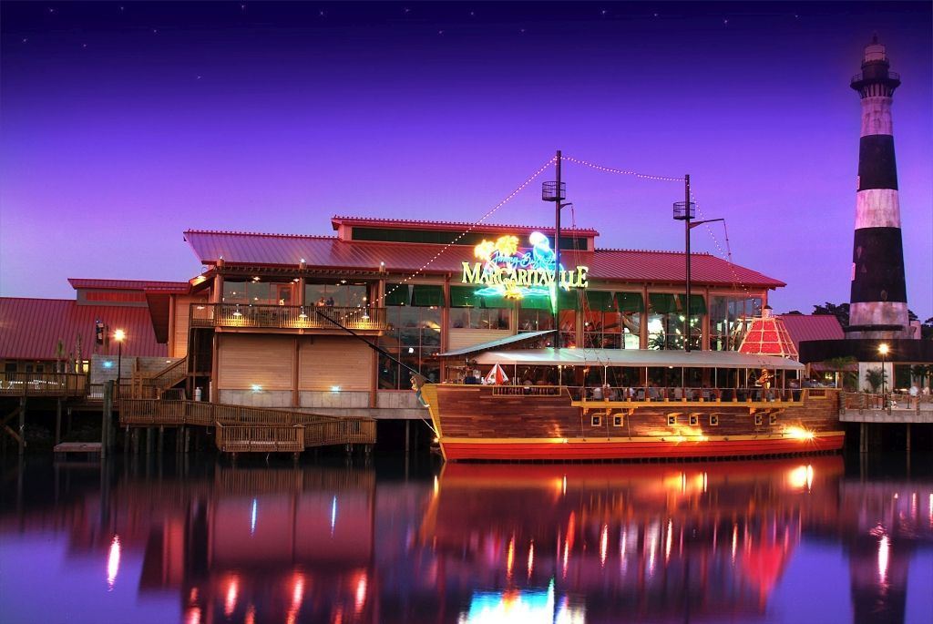 Restaurant and pirate ship on water at dusk, with lighthouse in background.