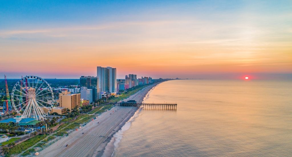 Beachfront at sunrise, with Ferris wheel and buildings. Ocean and colorful sky.