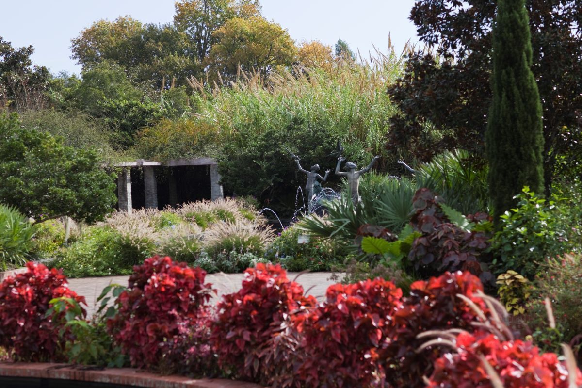 Garden scene with red plants in the foreground, a statue, and a covered structure.