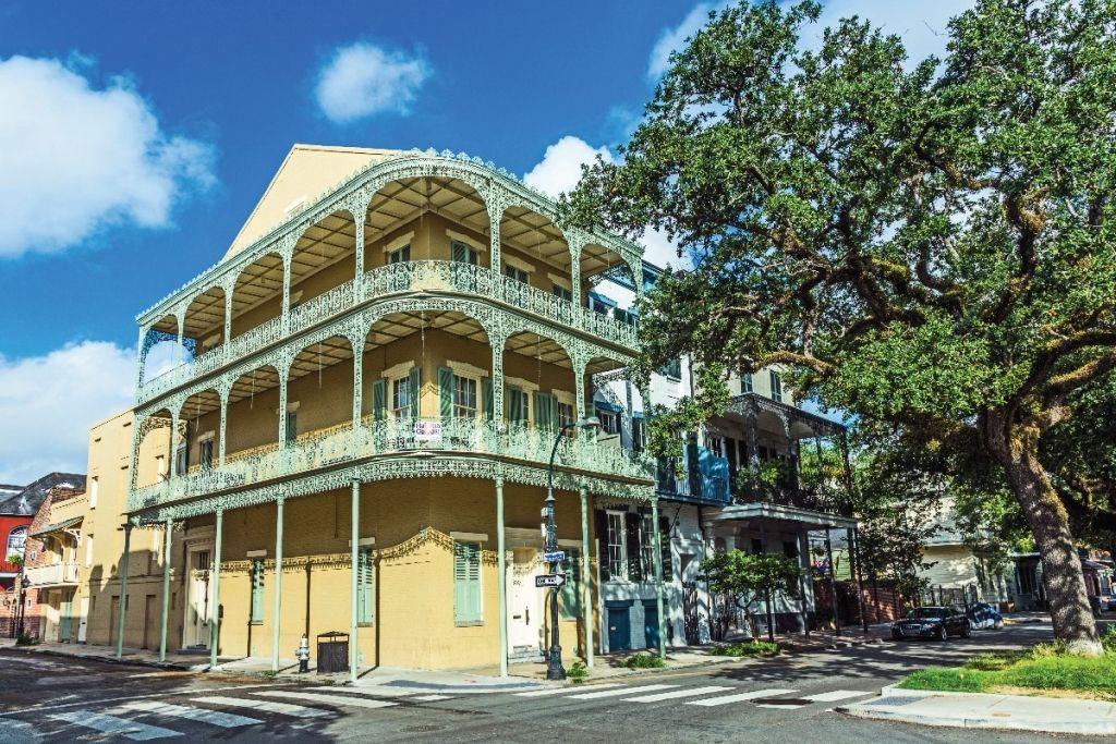 A yellow building with ornate wrought-iron balconies in New Orleans, with a large tree and blue sky.