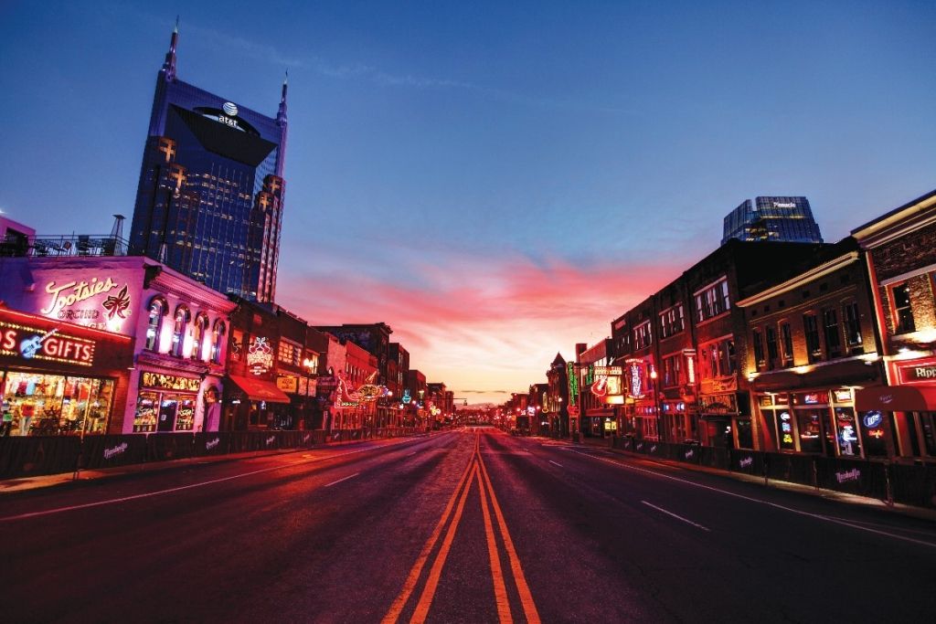 A street at dusk in Nashville, TN, lined with lit-up buildings under a pink and blue sky.
