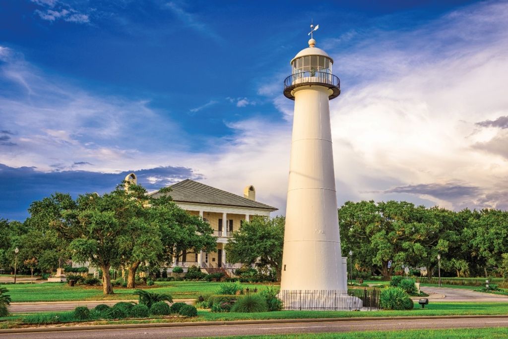 White lighthouse and mansion set against a blue sky with trees in foreground.