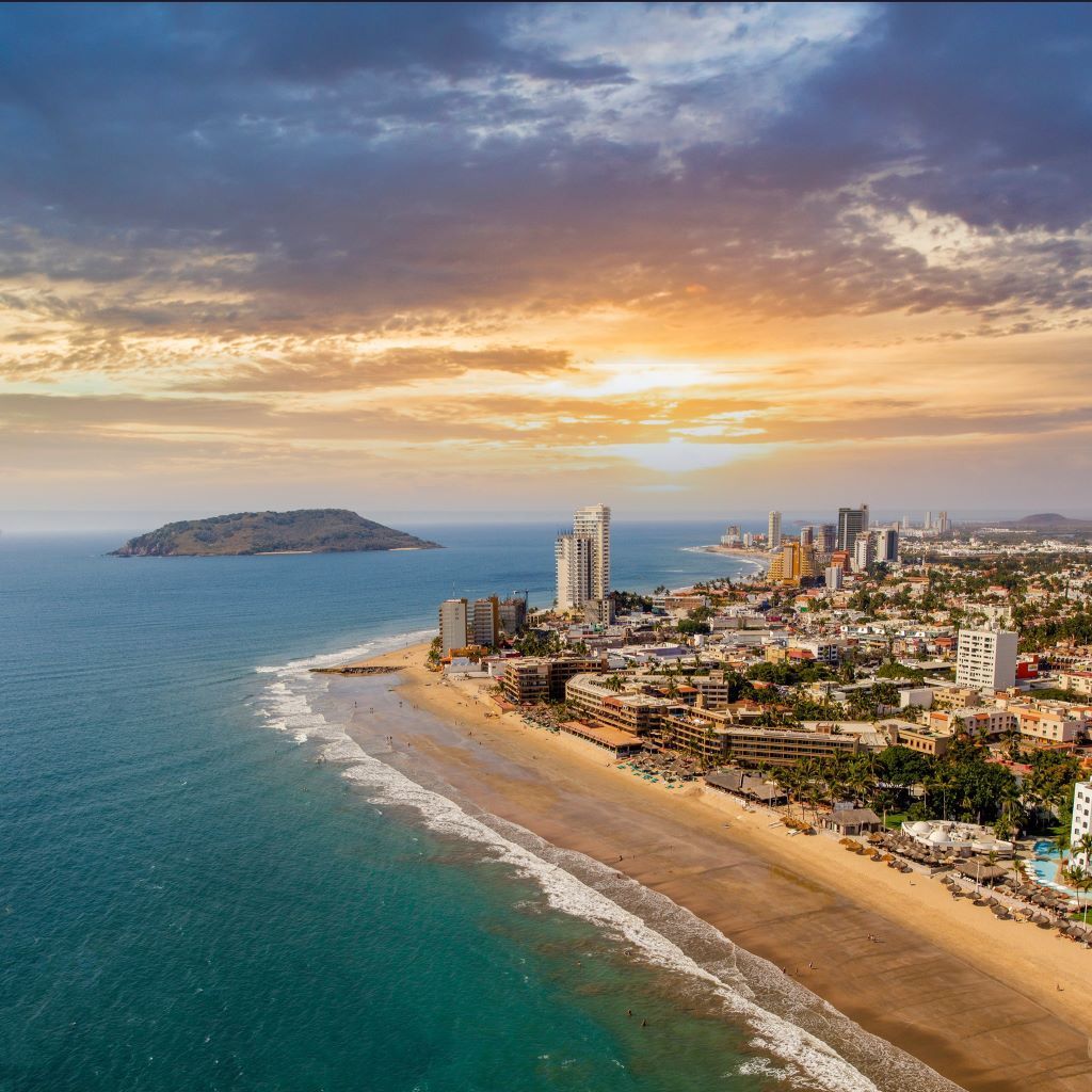 Aerial view of a coastal city with a sandy beach, turquoise sea, and sunset sky