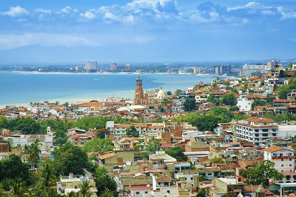 Coastal hillside cityscape with colorful buildings, green trees, and a blue bay under a clear sky