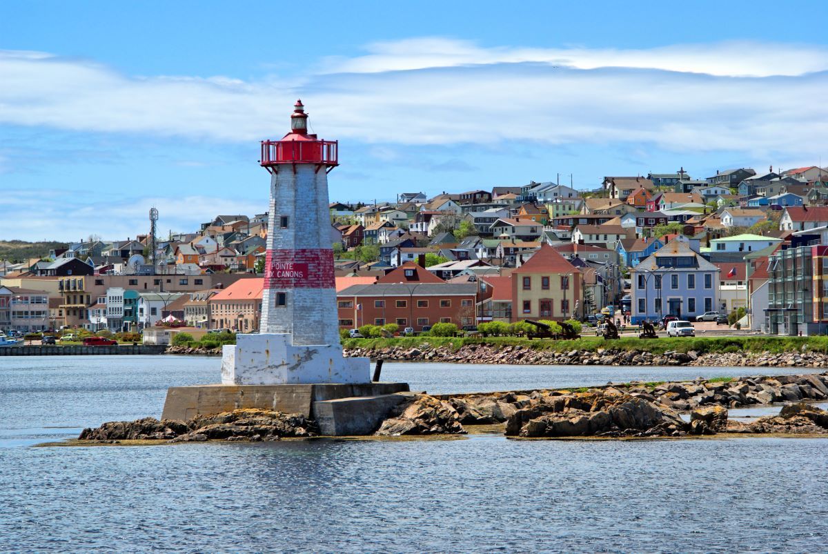 A white and red lighthouse stands on a rocky pier in front of a coastal town under a partly cloudy blue sky.