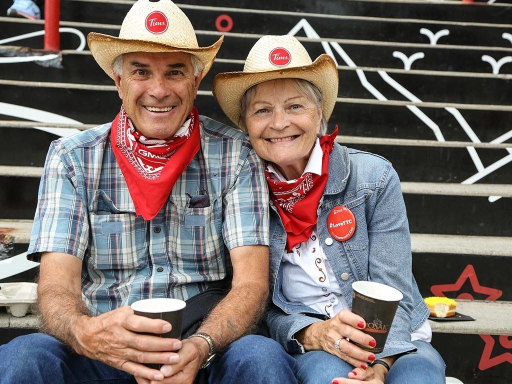 Smiling senior couple wearing cowboy hats and bandanas, holding coffee. Sitting on steps.
