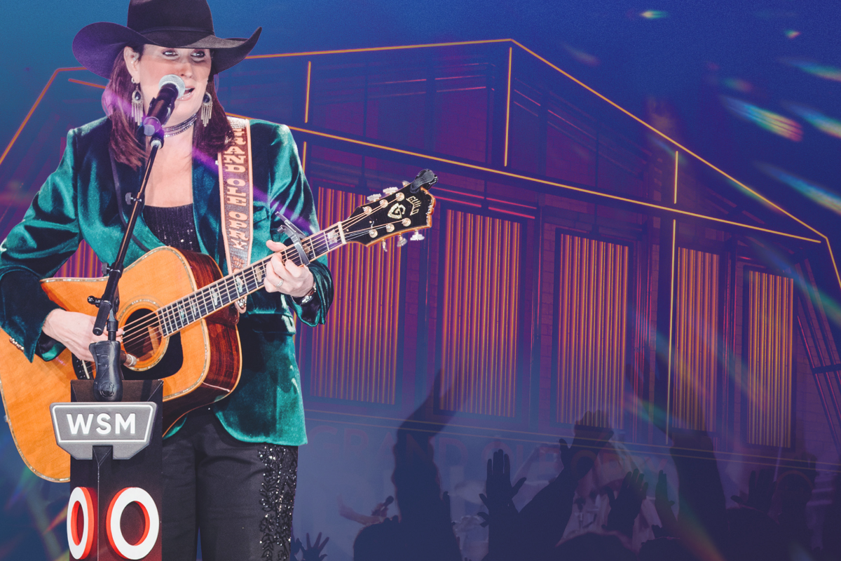 Singer in a cowboy hat playing acoustic guitar onstage with purple-lit backdrop, holding a microphone