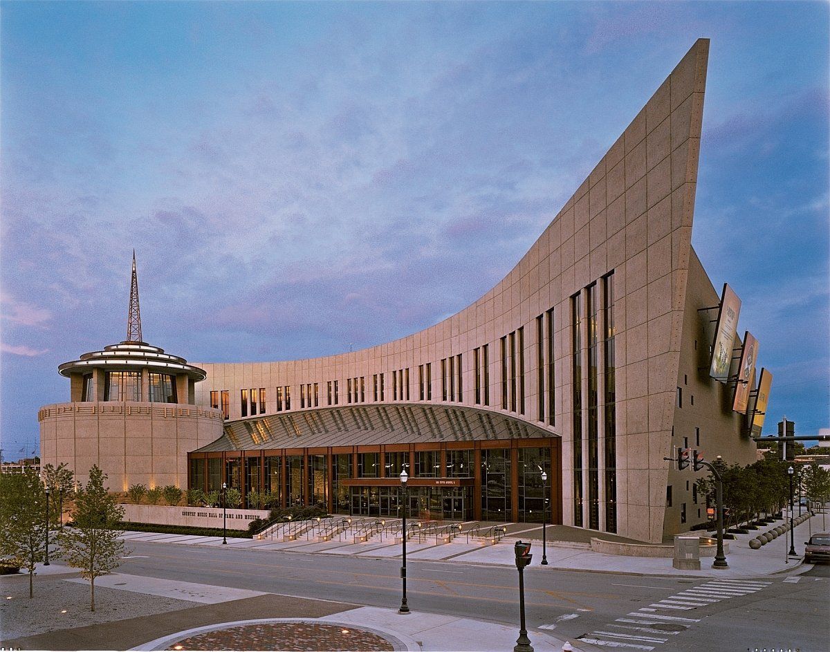Large, modern building with a curved facade and glass entrance. It has a spire and is near a street.