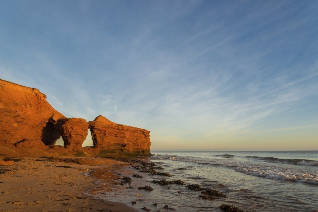 Red cliffs with archway on a sandy beach, waves and blue sky at sunset.
