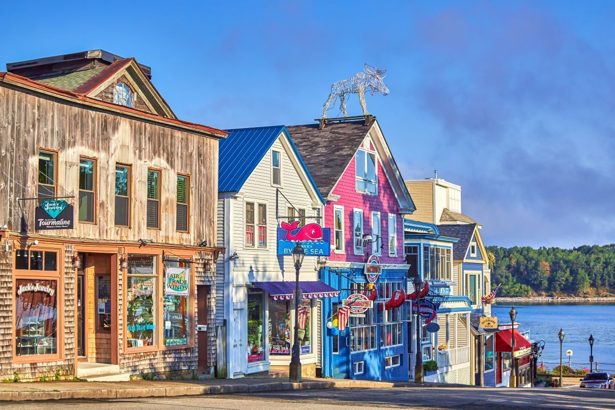 Colourful storefronts along a coastal road, blue sky, water in background.