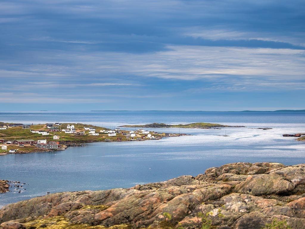Coastal village with white houses, blue water, and rocky foreground under a cloudy sky.