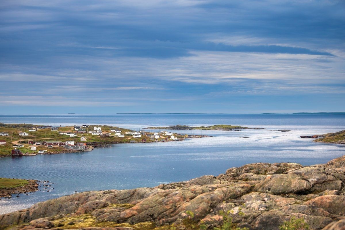 Coastal village with white houses and blue water under a cloudy sky, rocky foreground.