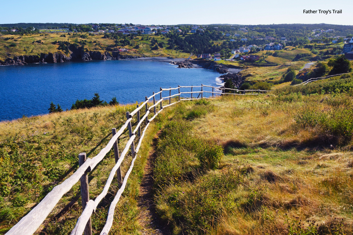 Coastal view with a wooden fence, path, blue water, green and brown grassy hills, and a town.