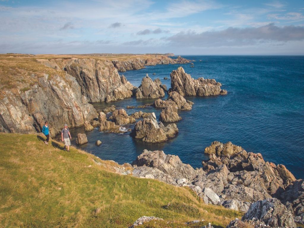 Cliffside coastal path overlooking rocky sea stacks and blue ocean, with a few hikers on the grassy slope