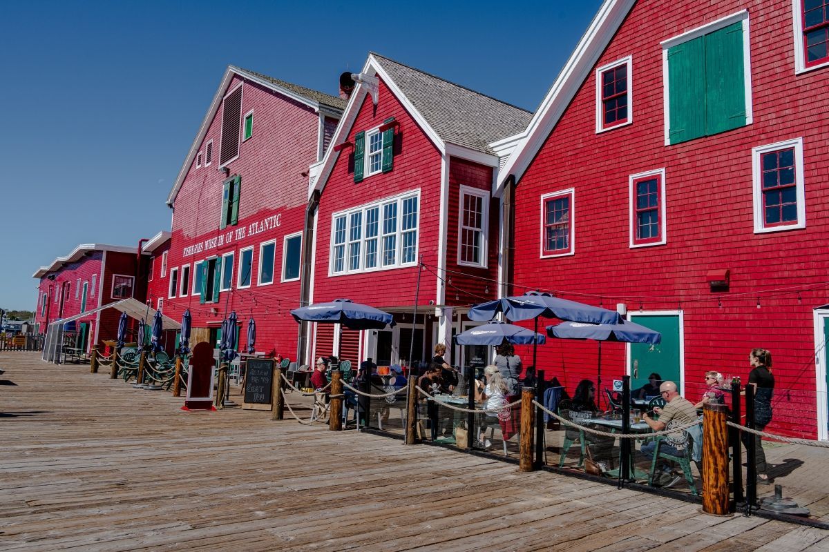 Red buildings with green shutters line a waterfront; outdoor tables with blue umbrellas.