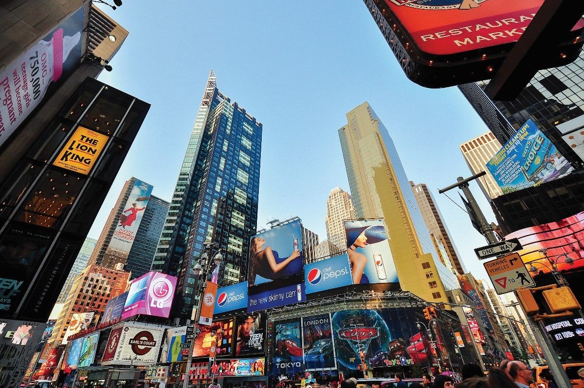 Times Square, NYC. Skyscrapers surround bright digital billboards. Blue sky overhead.