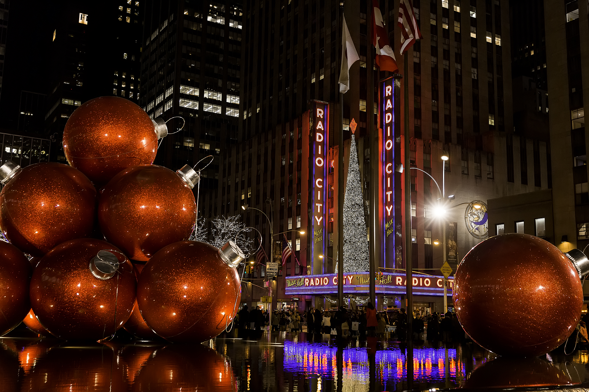 Giant orange Christmas ornaments in front of Radio City Music Hall and Christmas tree at night.