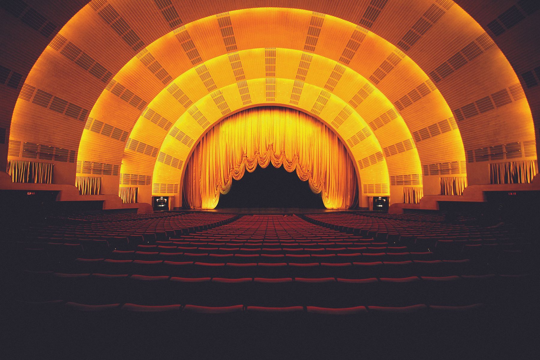 Interior view of a theater stage lit with warm yellow lights, plush seating, and a closed golden curtain.