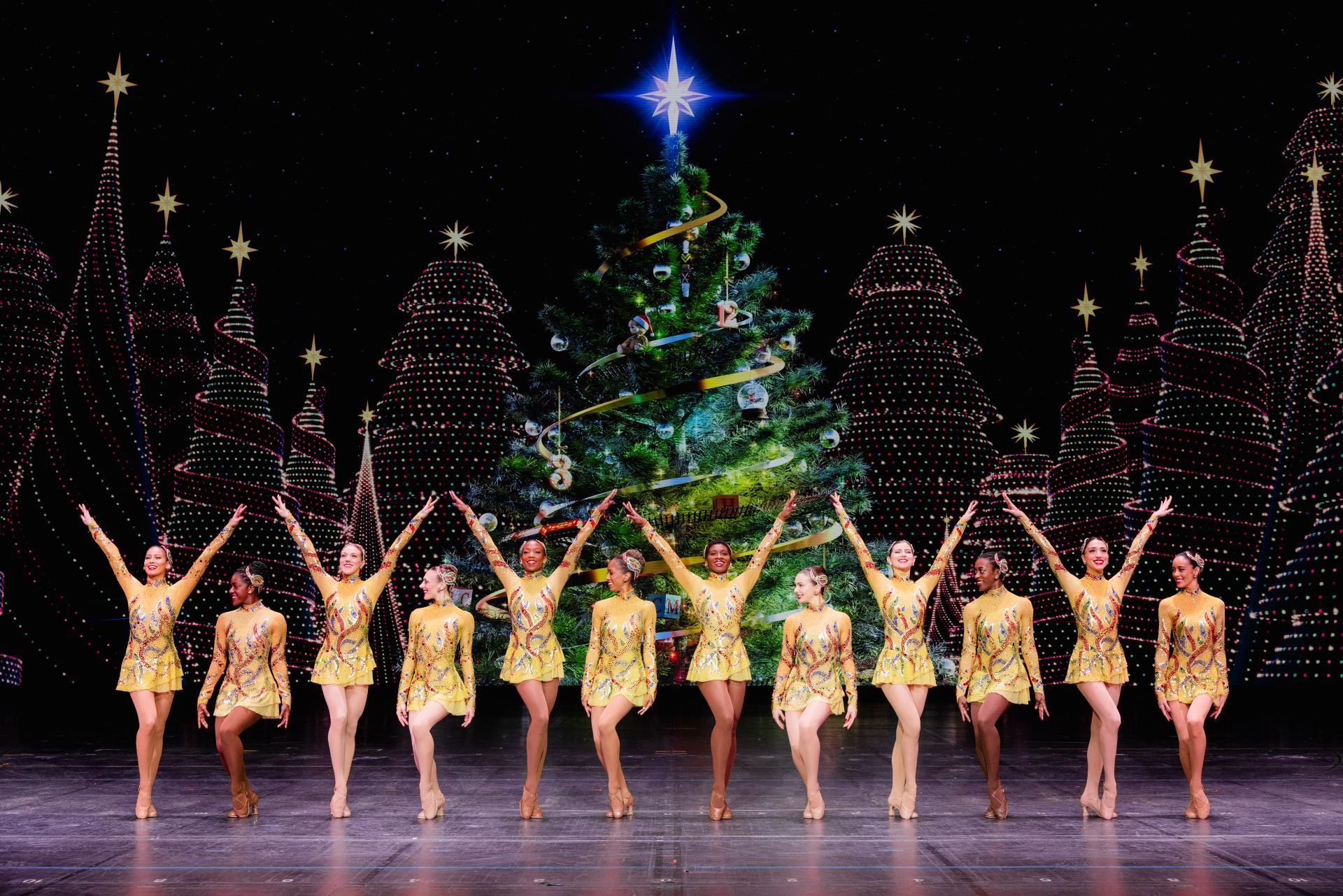 Dancers in gold costumes with arms up, in front of a Christmas tree stage set.