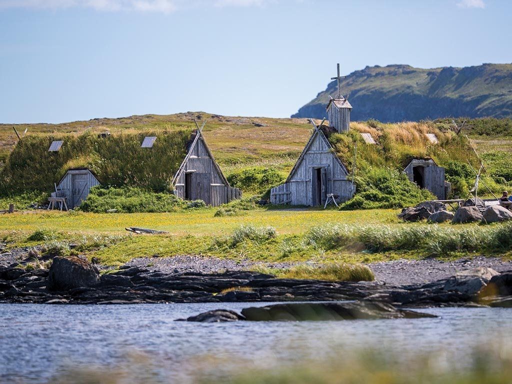 Grassy-roofed wooden buildings near water, with a lighthouse in the background under a blue sky.