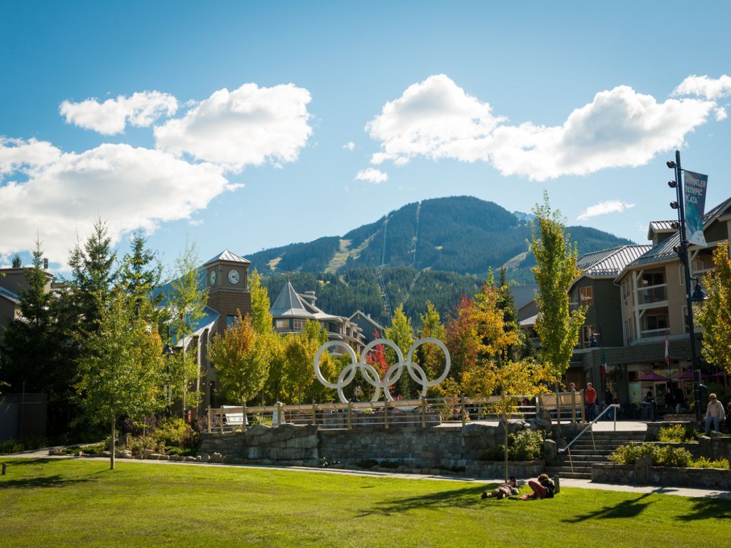 Olympic rings in park with mountains in background. Buildings, trees, and blue sky.