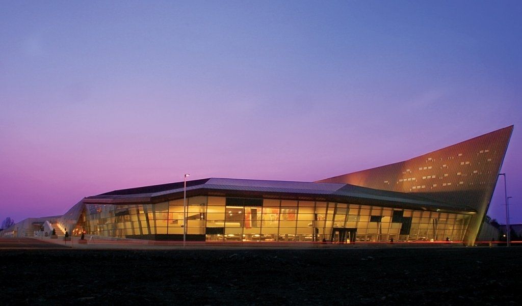 Modern building at dusk with angular, glass facade and golden roof against a purple sky.