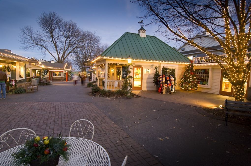 A festive outdoor shopping area at dusk, with shops, Christmas lights, and a table with a floral centerpiece.