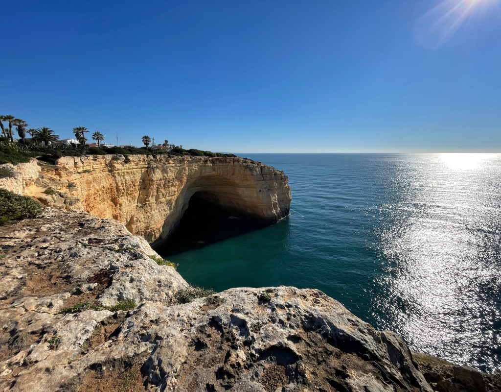 Cliffside cave overlooking the ocean under a bright blue sky. Sunlight reflects on the water.