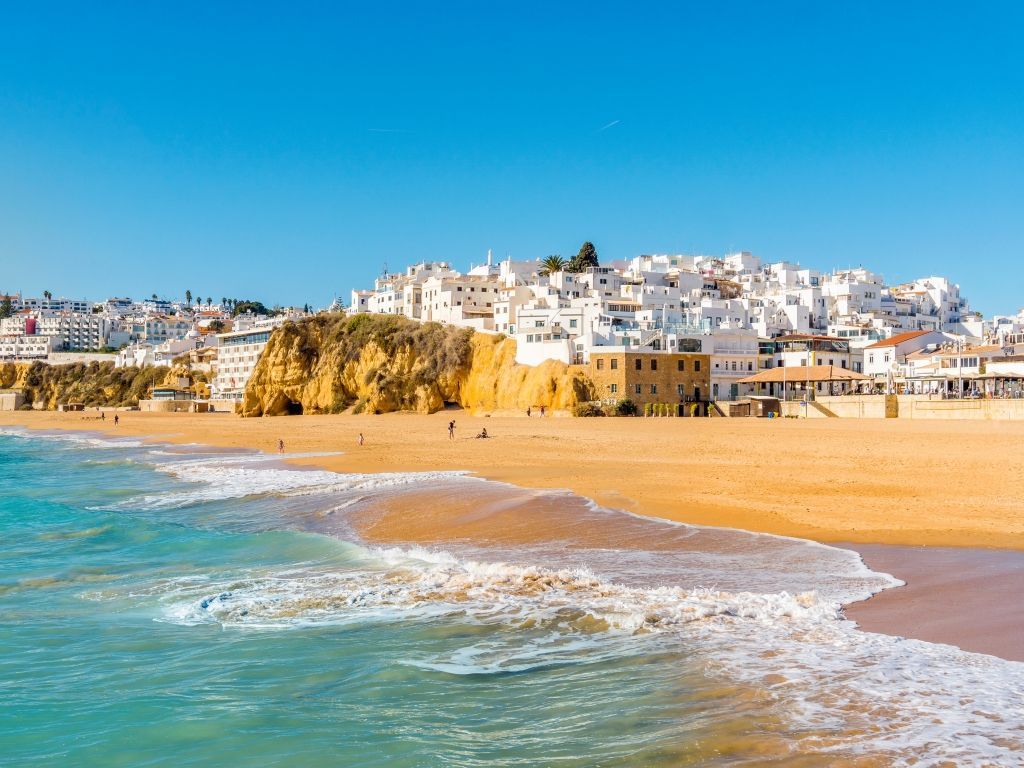 Sunny beach with turquoise waves and white hillside buildings under a clear blue sky