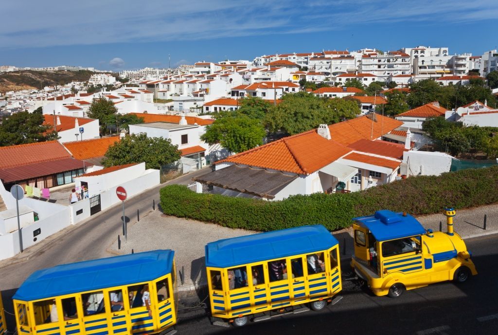Yellow and blue tourist train traveling past white buildings with red tile roofs.