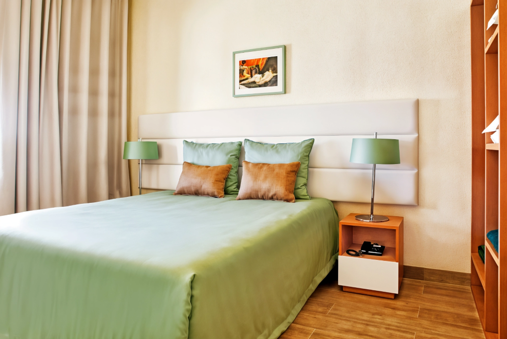 Neatly made bed with green bedding in a bright bedroom, flanked by matching bedside lamps.