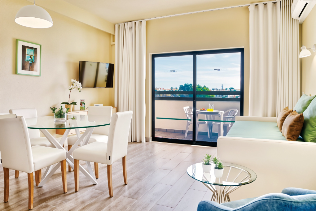 Bright modern living-dining room with balcony, glass table, white chairs, and sofa by large windows