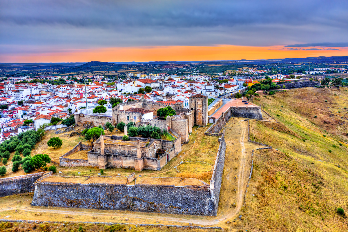 Aerial view of a fortress overlooking a white-roofed town at sunset. Yellow grass and stone walls.