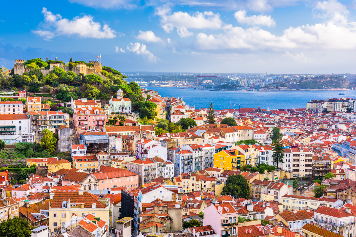 Cityscape of Lisbon, Portugal, with red-tiled roofs, buildings, and a hilltop castle overlooking the Tagus River.