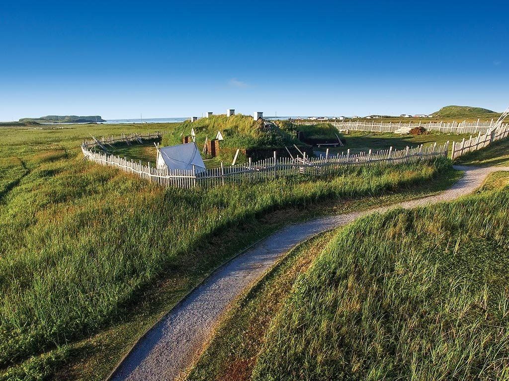 Historic Viking settlement with sod-covered structures inside a wooden palisade fence on grassy land under a blue sky.