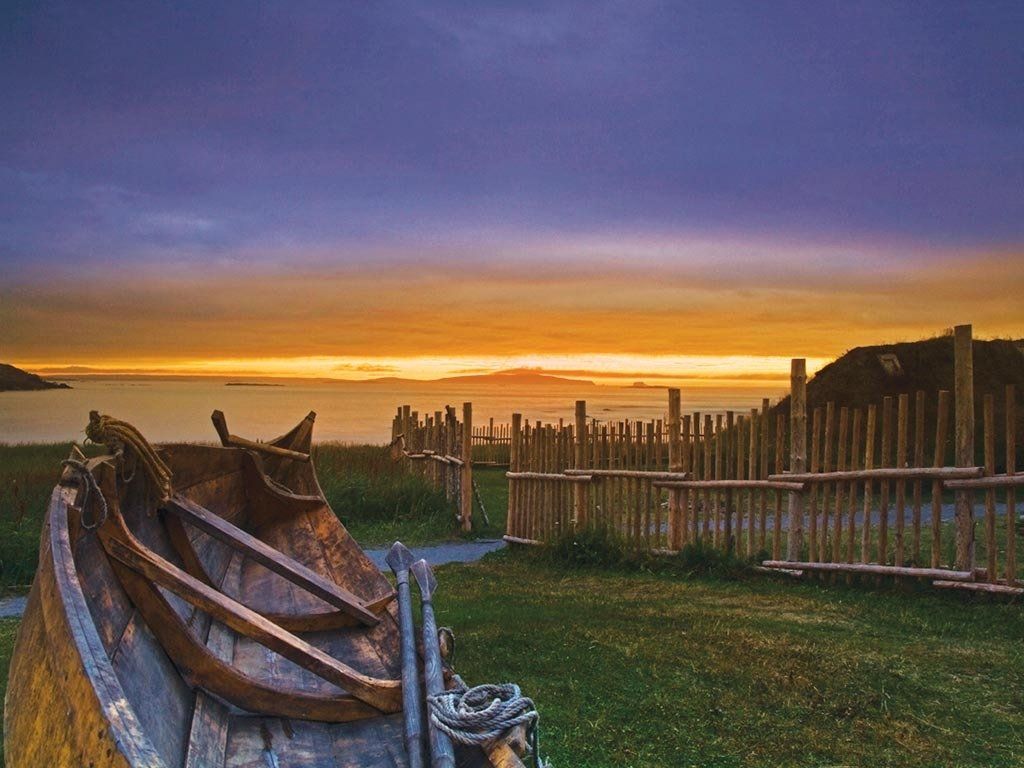 Old wooden boat on grass overlooking ocean at sunset, with fence and dark sky.