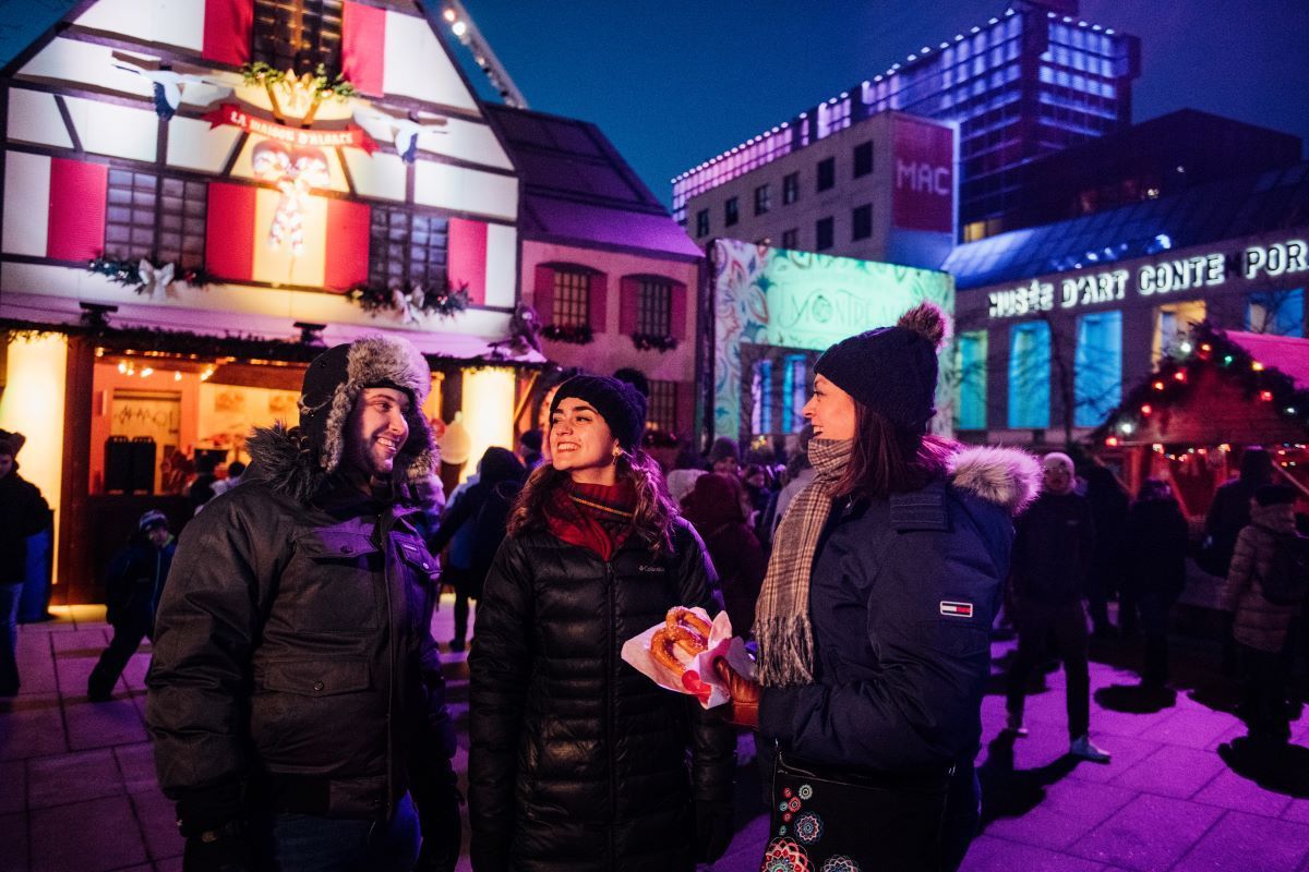 Three friends at a winter market, smiling, dressed in warm clothes, colorful lights in the background.