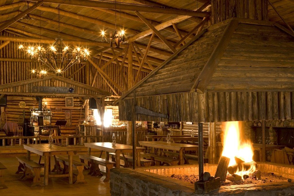 Rustic lodge interior with wooden tables, benches, and a large fireplace, bathed in warm light.