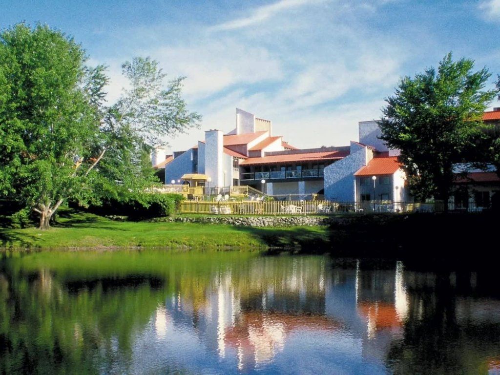 Lakeside resort with white buildings, red roofs, and trees reflected in the calm water. Blue sky overhead.