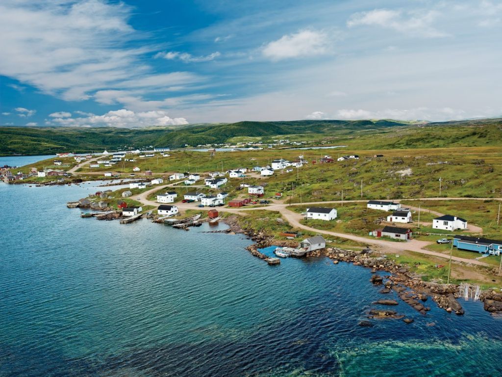 Coastal village with white houses, blue ocean, green hills under a sunny sky.