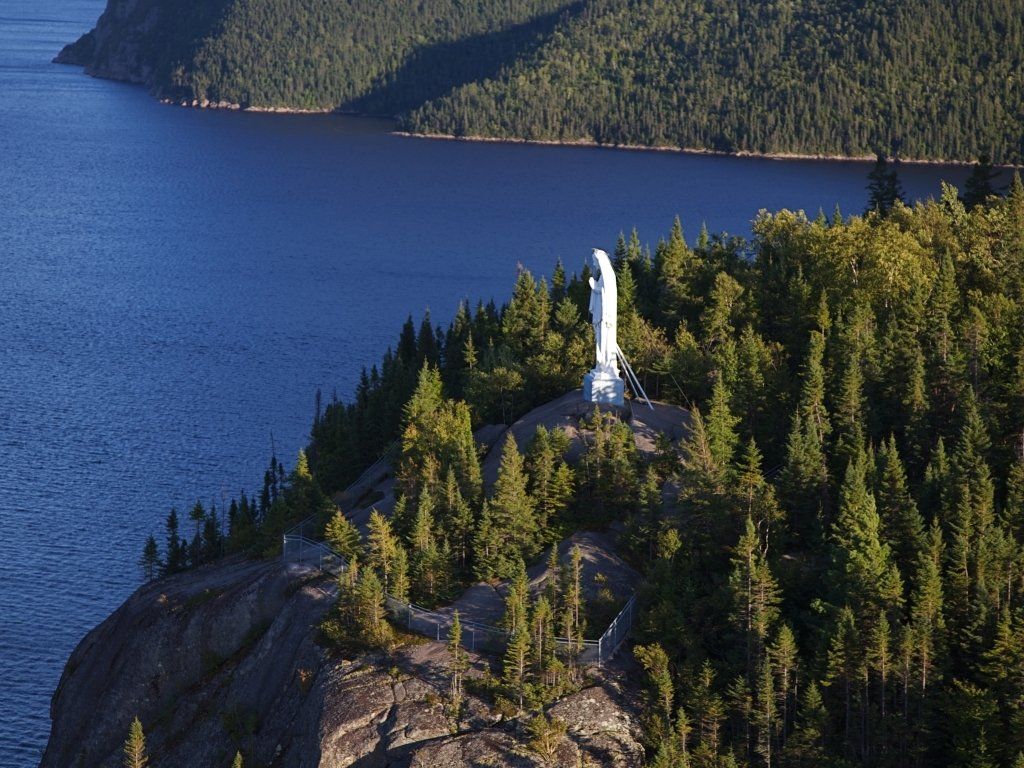Statue of the Virgin Mary atop a forested cliff overlooking a deep blue lake.